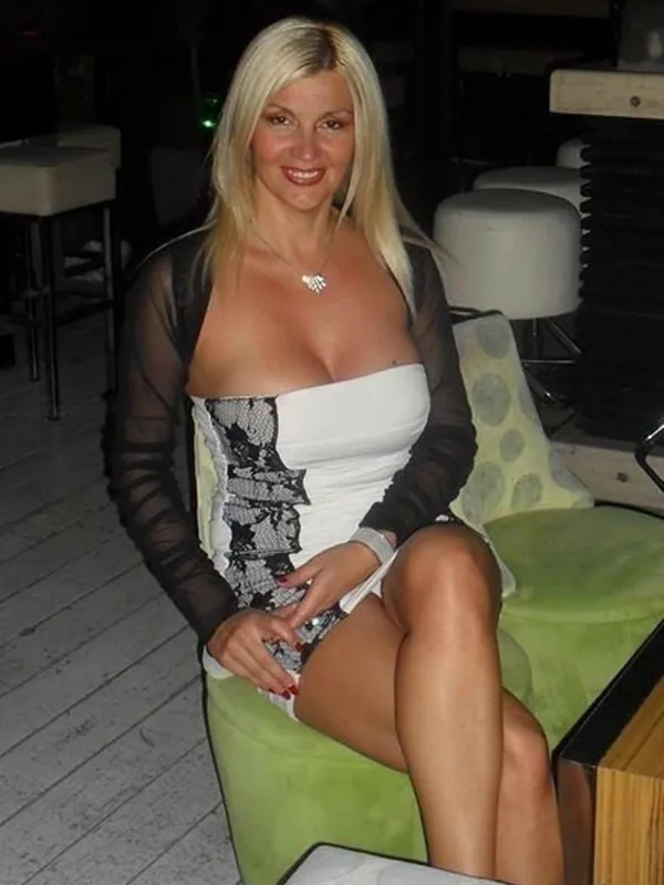 Femme blonde assise sur un fauteuil vert, portant une robe blanche et une veste transparente noire, souriant à l'appareil.