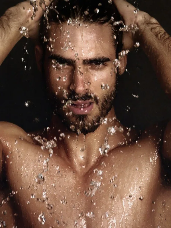 Homme torse nu sous une pluie d'eau, les mains dans les cheveux, regardant l'objectif.