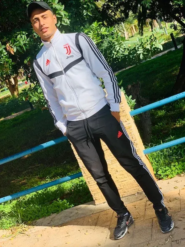 Homme en survêtement Juventus posant dans un parc ensoleillé, appuyé sur une barrière bleue.