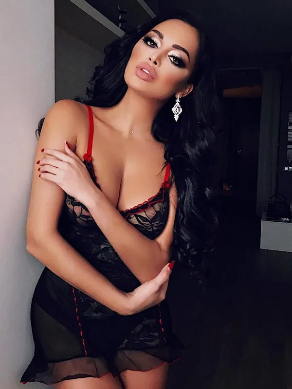Femme aux cheveux noirs portant une lingerie noire à dentelle avec des détails rouges, posant près d'un mur dans un intérieur sombre.
