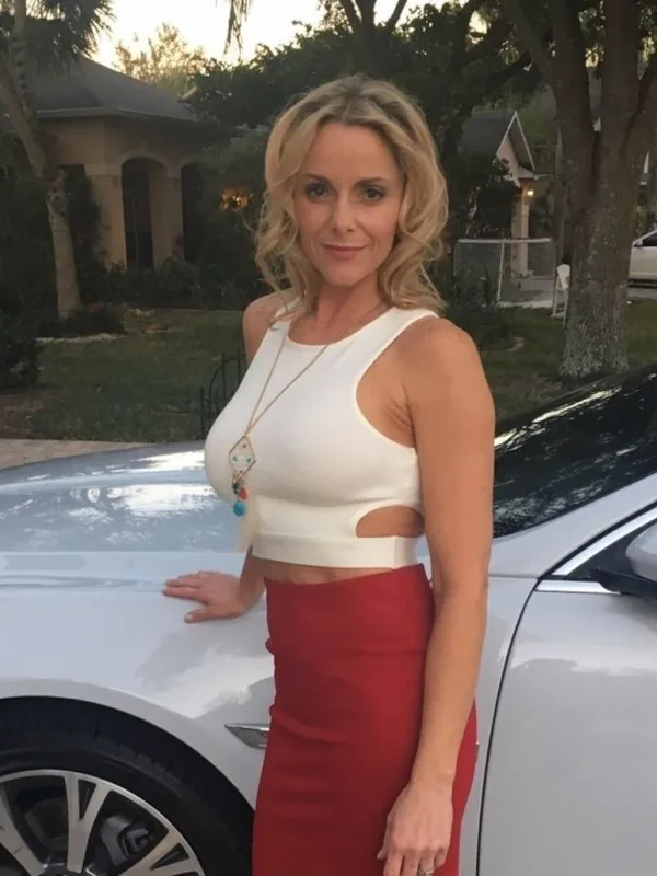 Femme blonde en haut blanc et jupe rouge posant devant une voiture blanche dans un jardin au crépuscule.